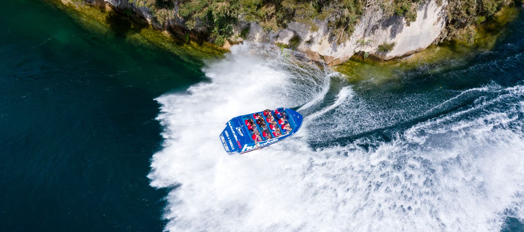  The image shows an aerial view of a blue jet boat labeled "hukafalls" making a sharp turn on a clear, greenish-blue river. The boat, filled with passengers wearing red life jackets, creates a large white wake as it maneuvers near the rocky riverbank line