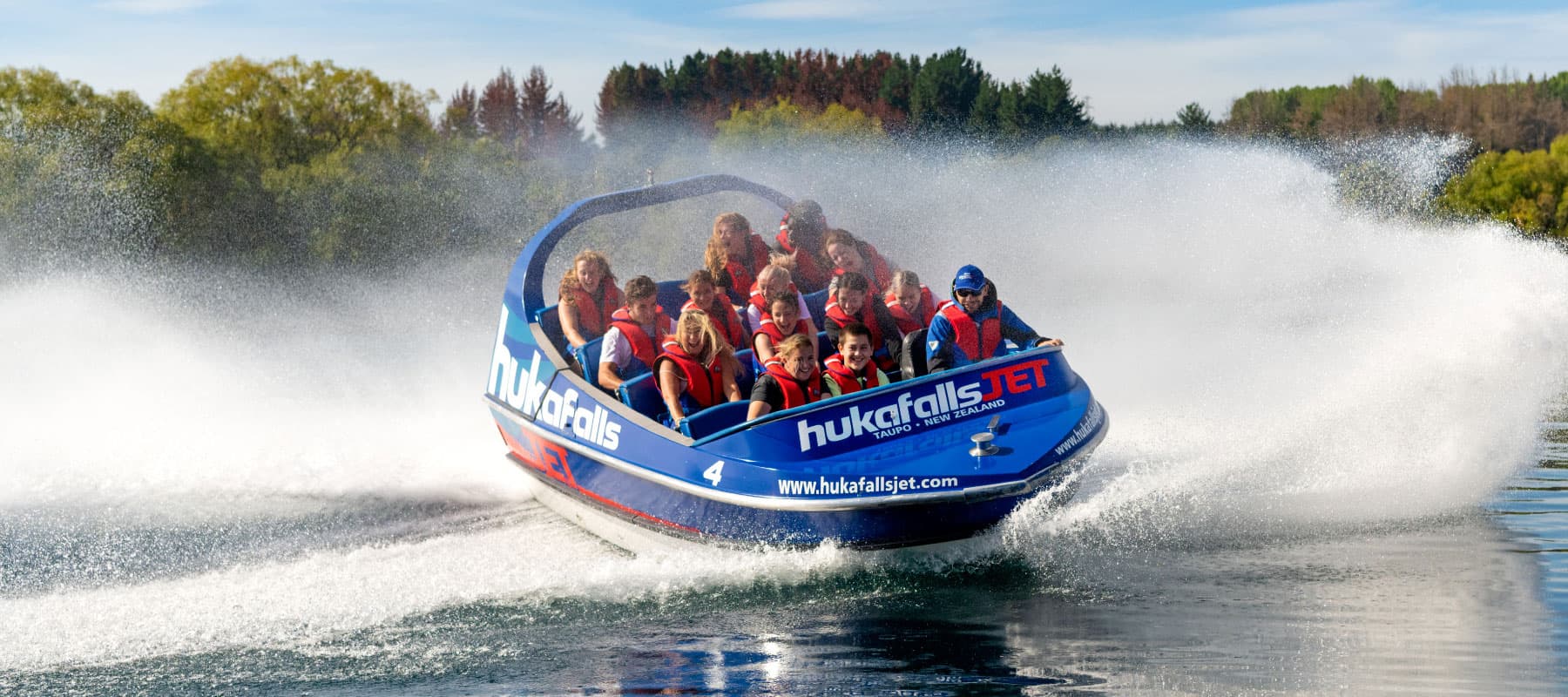  The image shows a blue jet boat labeled "hukafalls JET" speeding across the water, with passengers wearing red life jackets. The boat creates a large spray of water as it turns, and the passengers appear to be enjoying the ride, smiling and holding on ti