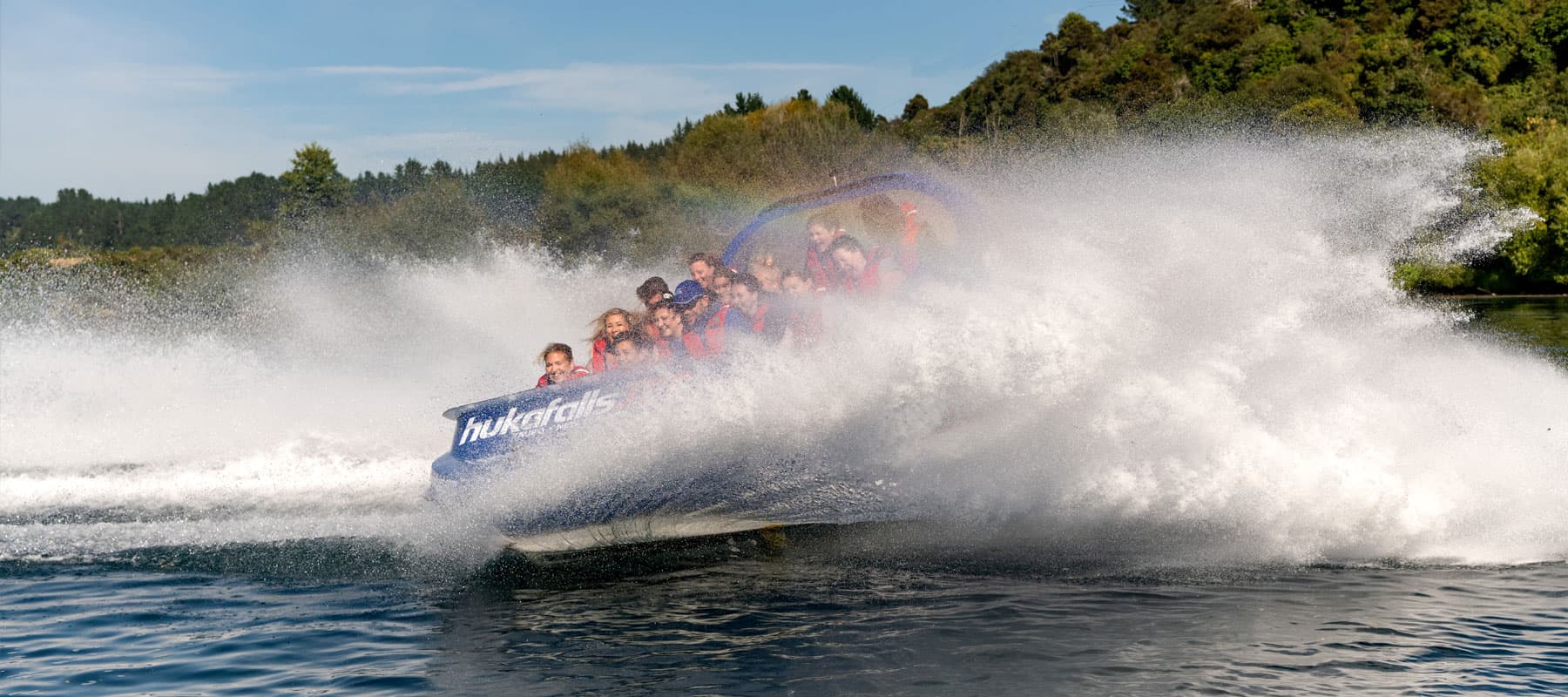 The image shows a blue jet boat labeled "hukafalls JET" speeding across the water, creating a massive splash of water that engulfs the boat. The passengers, wearing red life jackets, are seen smiling and laughing as they experience the thrilling ride. The